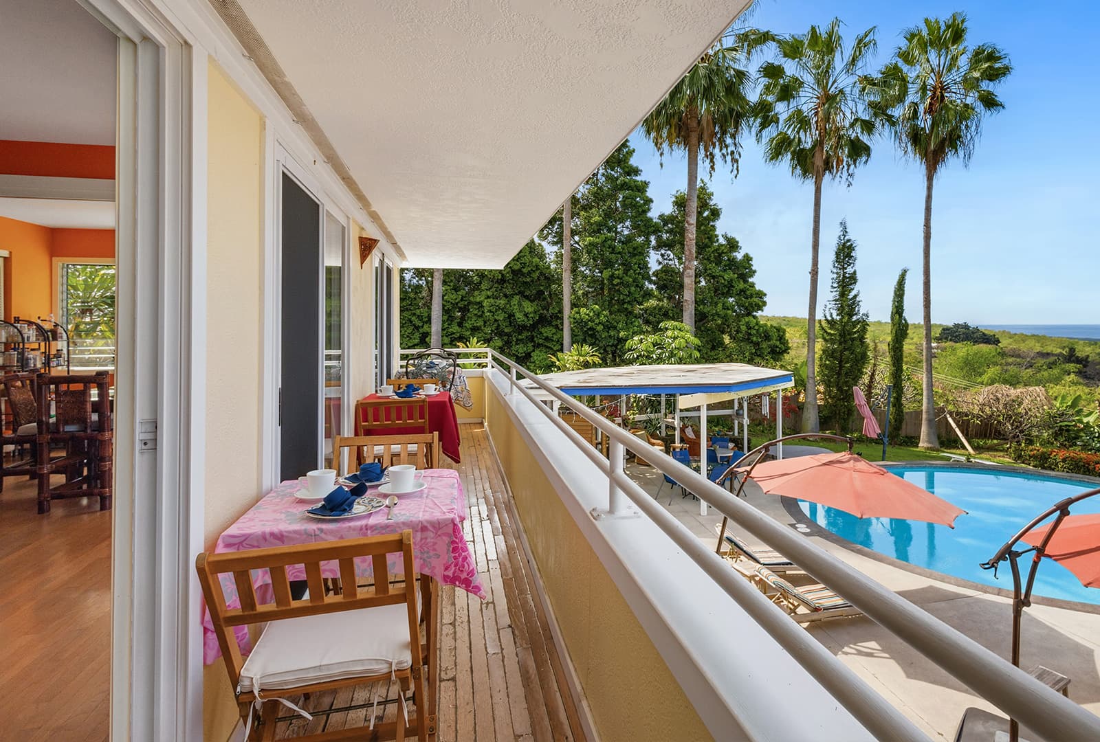 A sunny balcony with tables set for breakfast overlooks a pool surrounded by palm trees and greenery.
